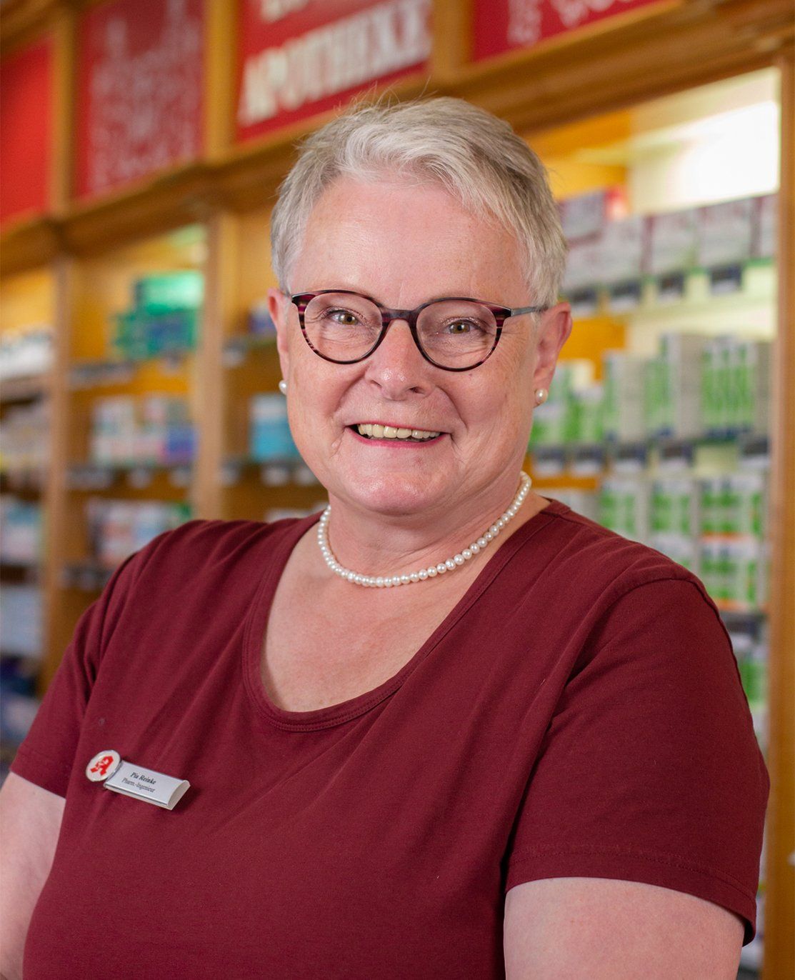 Eine Frau mit Brille und Perlenkette lächelt in einer Apotheke.