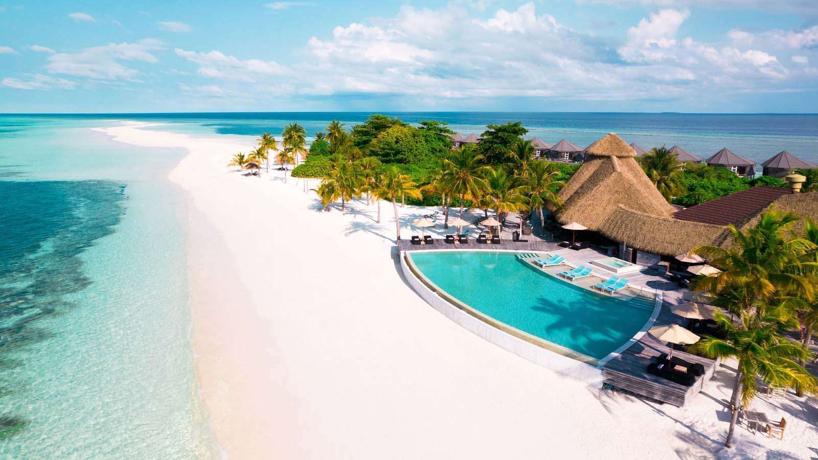 Tropical island resort with white sand beach, turquoise pool, and thatched roof buildings.