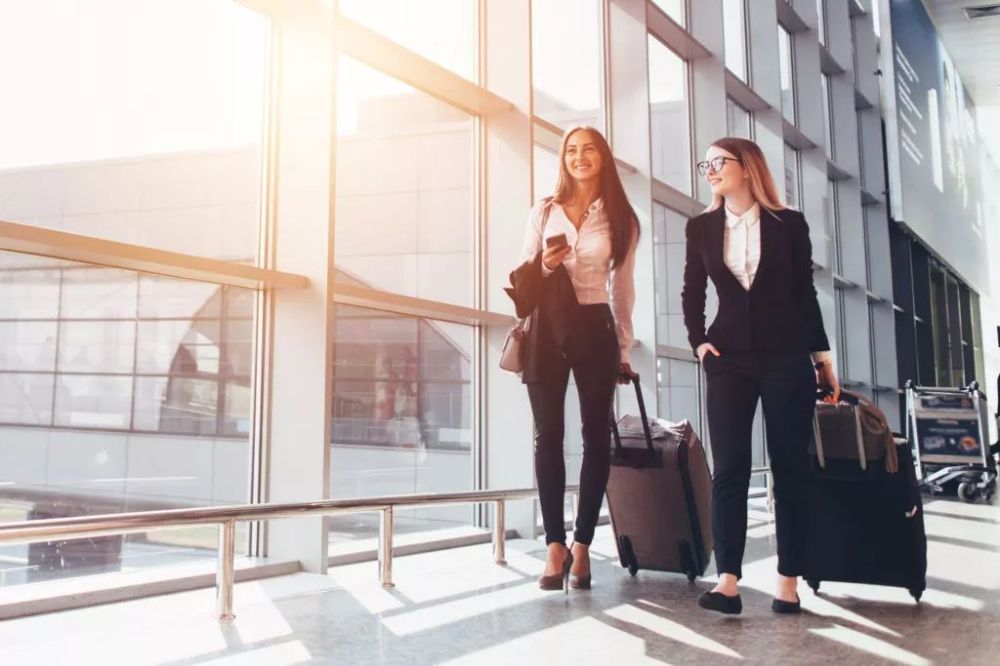 Two women in business attire walking through an airport, pulling luggage.