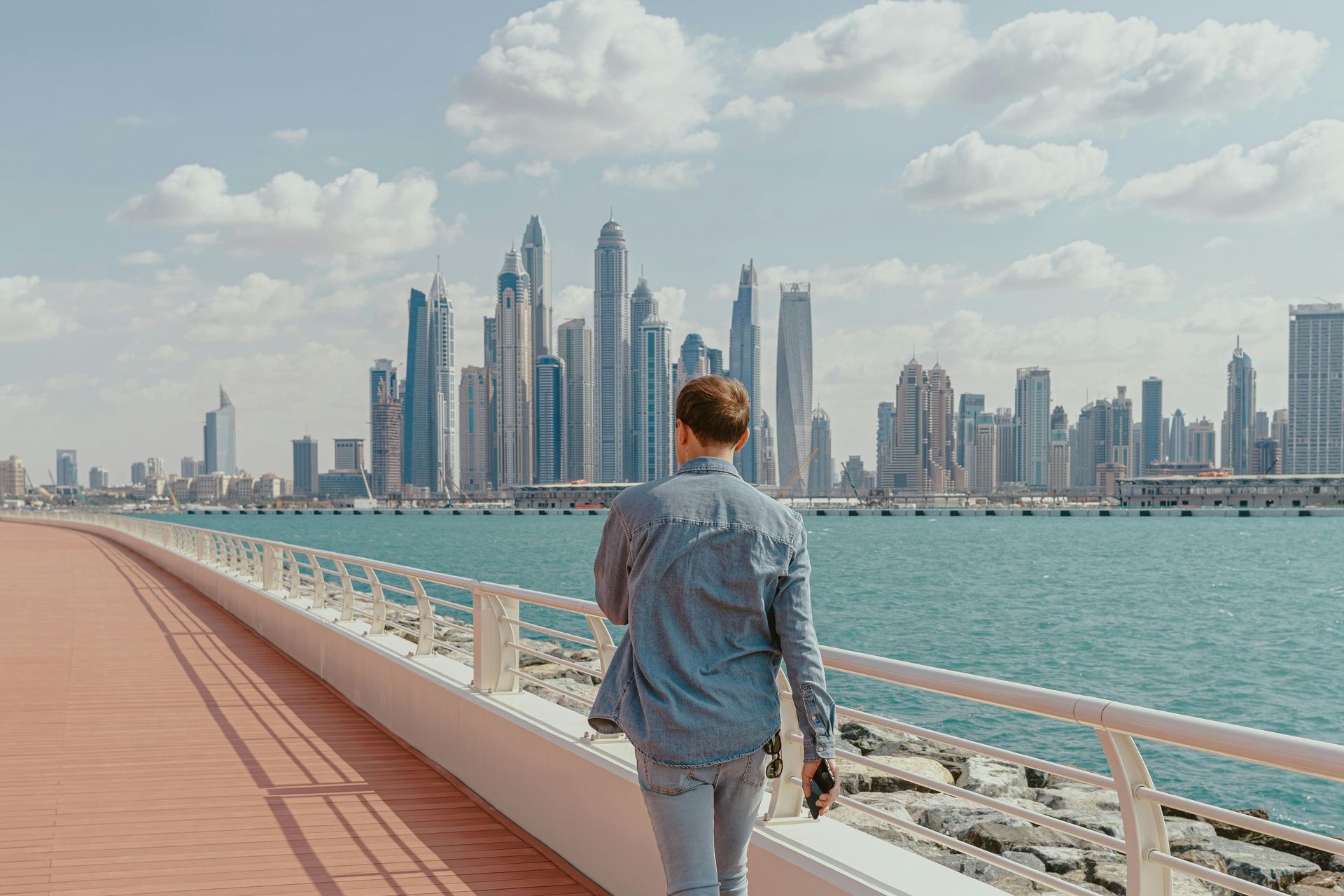 Person walking along a waterfront promenade, with a modern city skyline in the background.
