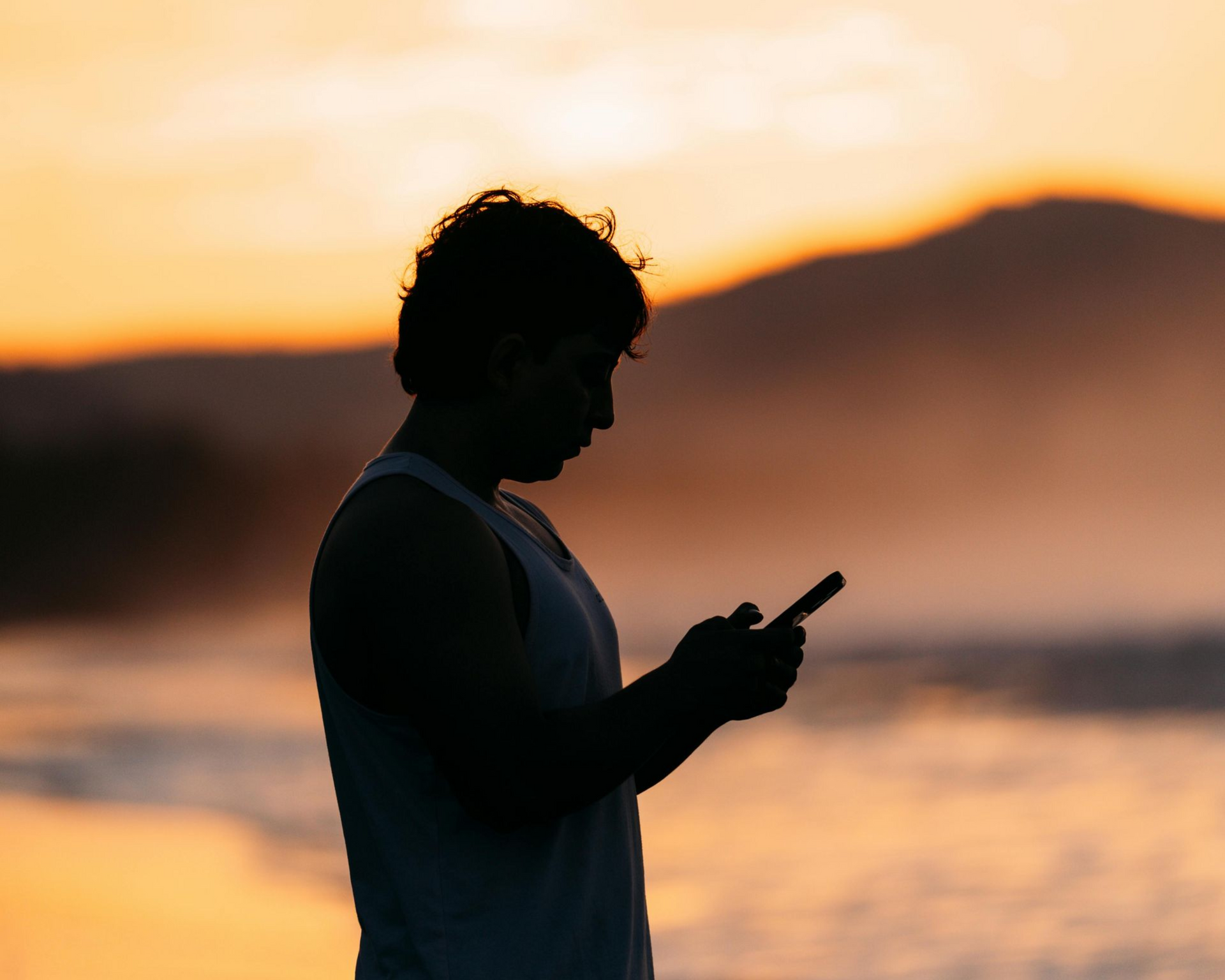 Silhouette of a person on a beach looking at a phone, silhouetted against a sunset over water and mountains.