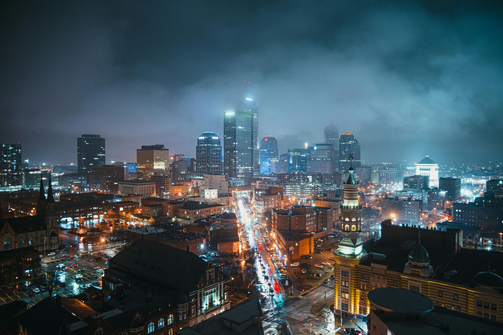 Nighttime cityscape with illuminated buildings, streetlights, and a cloudy sky.