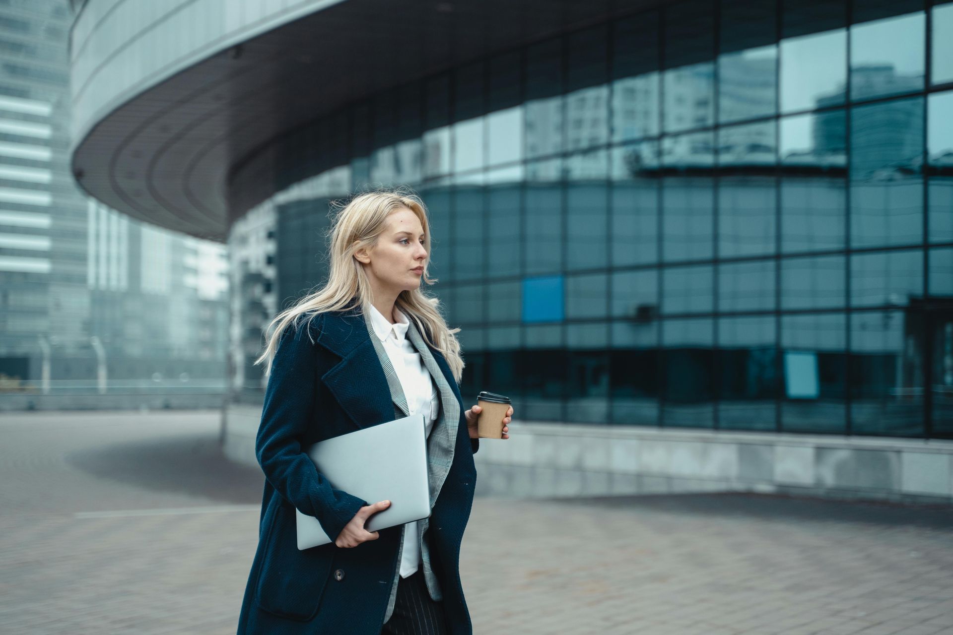 Blonde woman in navy coat, holding laptop and coffee, walking outside a modern building.