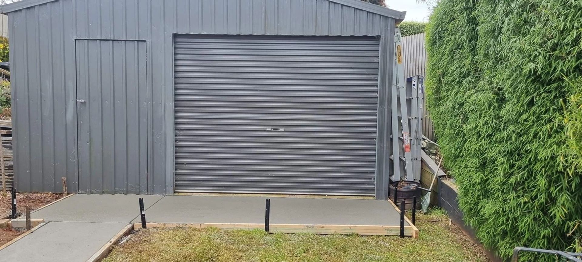 A gray shed with a roll-up garage door and a side door on a concrete pad. Lush green hedge on the right.