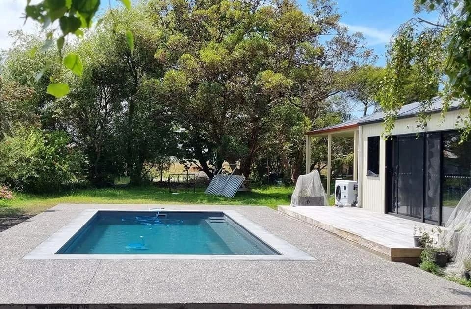 A small, rectangular outdoor pool with blue water, surrounded by a stone patio and greenery, next to a small building.