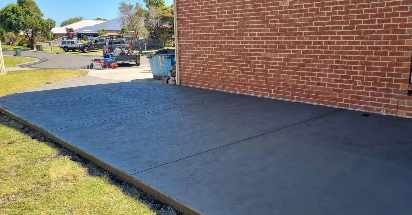 Newly poured dark gray concrete driveway next to a red brick building and grassy area on a sunny day.