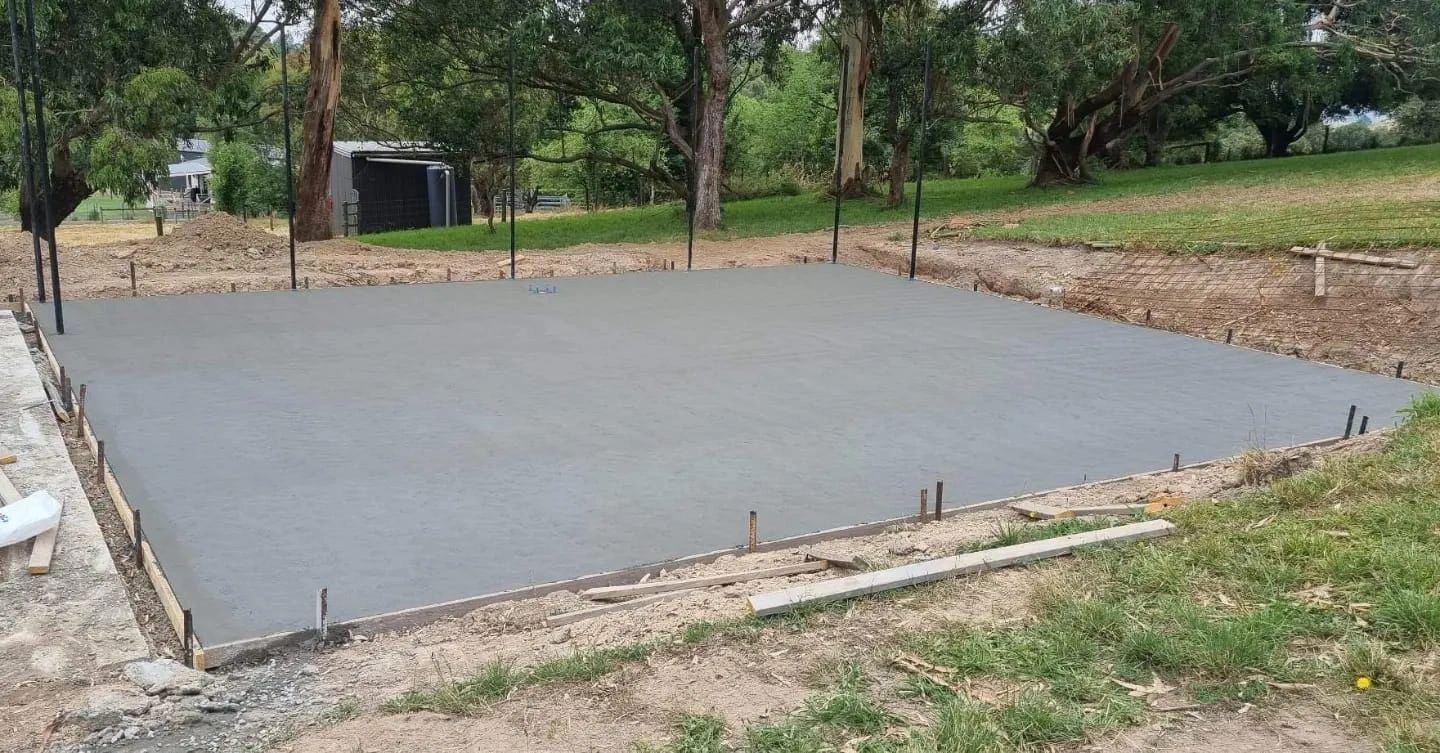 A freshly poured concrete slab, gray in color, framed by wooden boards, sits in a grassy yard with trees in the background.