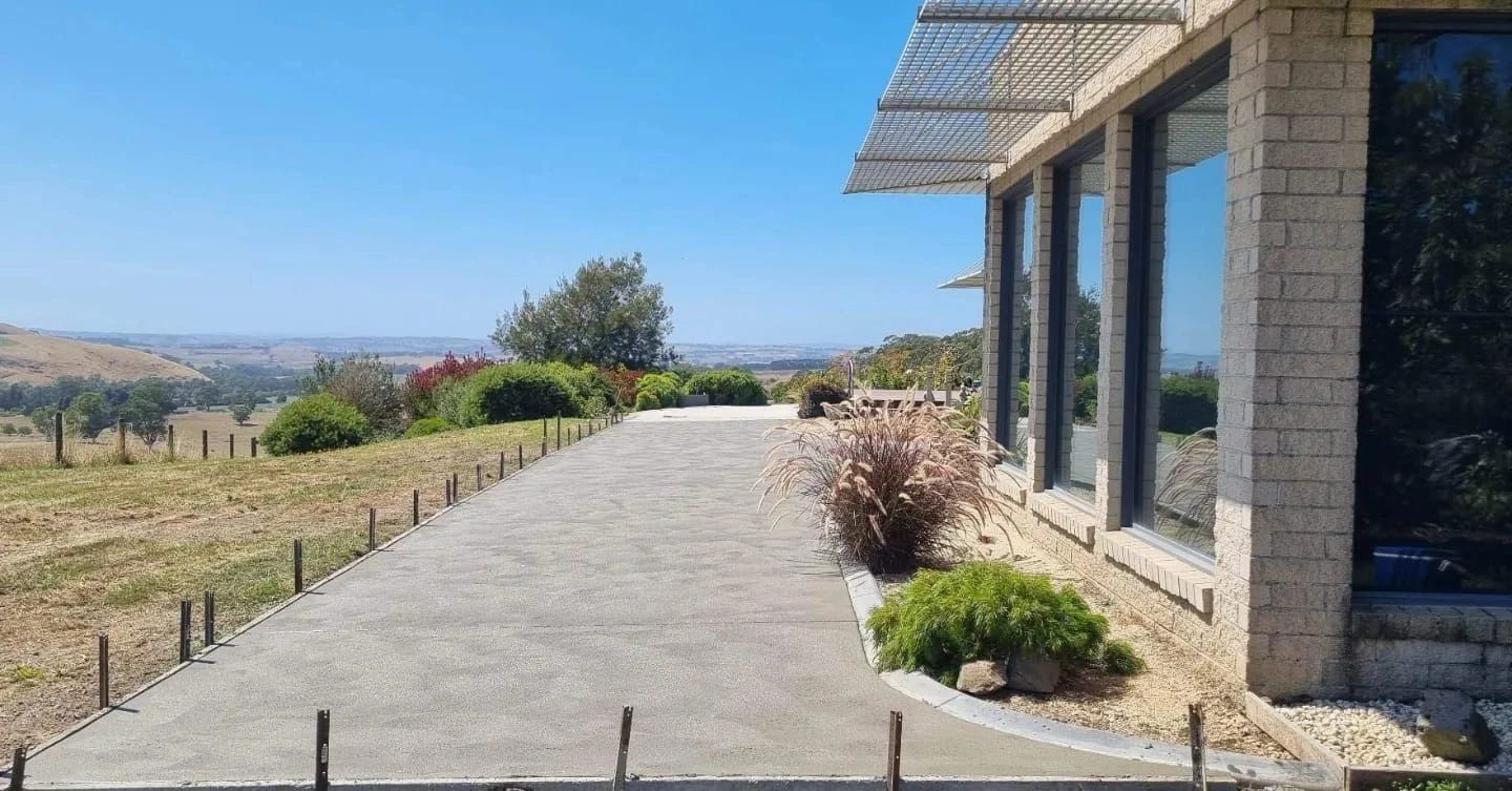 A long concrete pathway next to a stone-walled building with large windows. The pathway is bordered by metal posts, leading to a distant landscape under a blue sky.