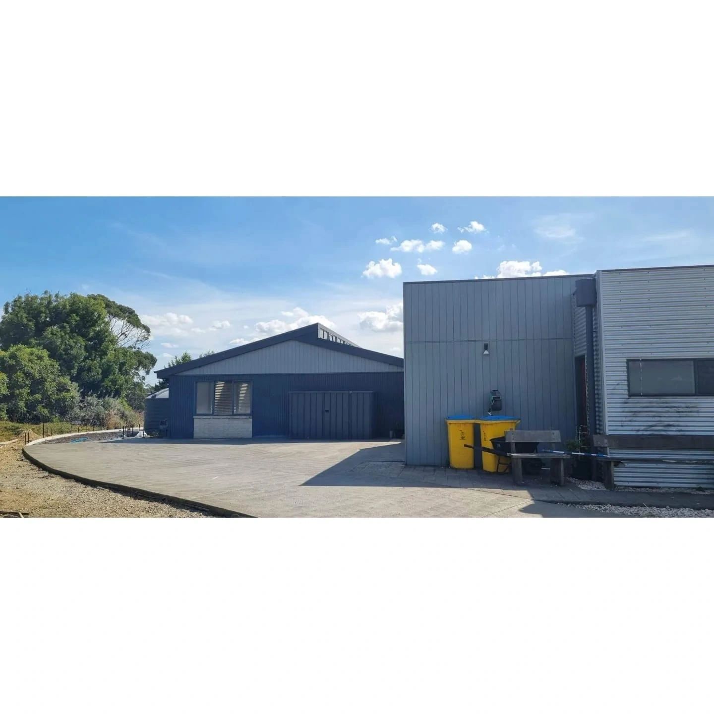 Blue industrial buildings with a paved area and yellow bins against a blue sky. Trees are visible to the left.