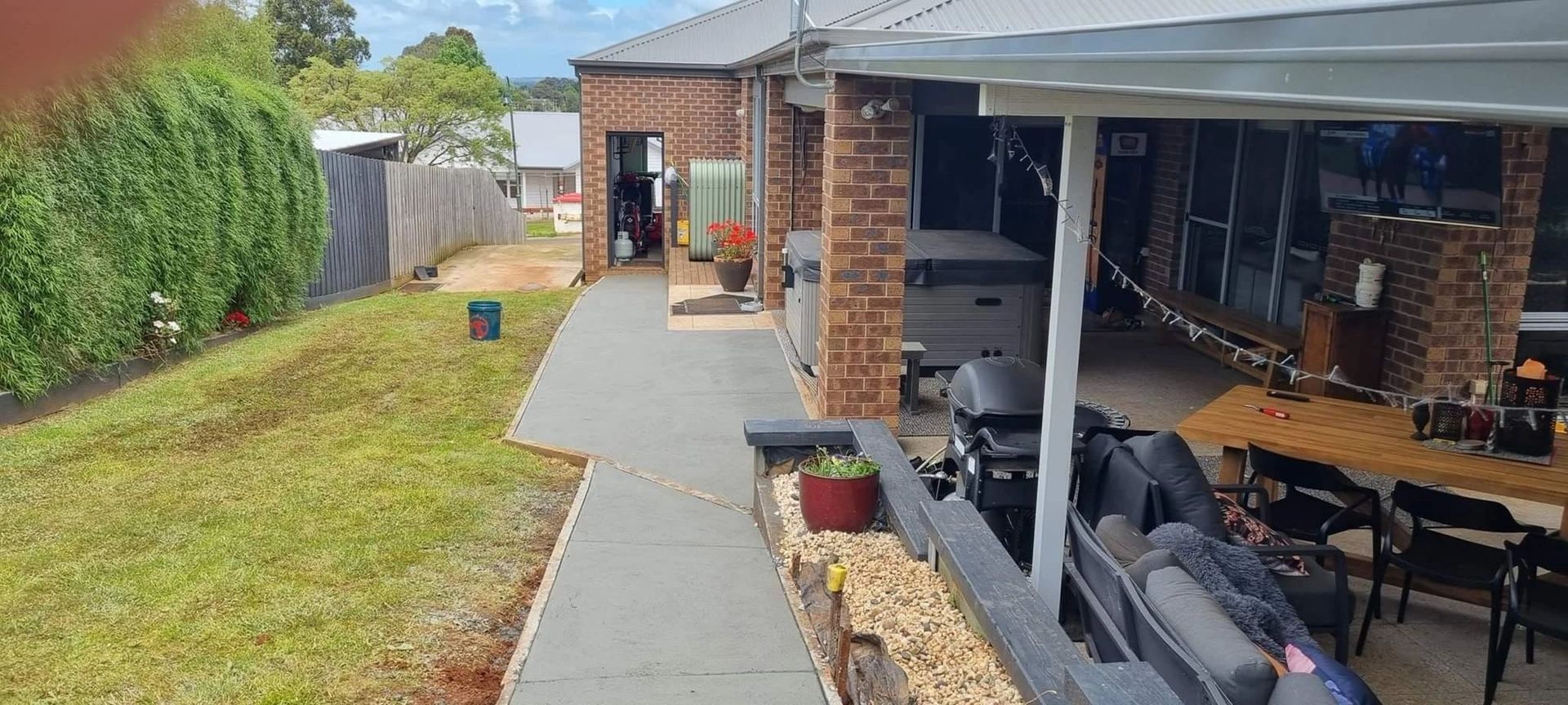 Backyard view with a covered patio, hot tub, and outdoor furniture. A walkway leads past a brick home and grass.