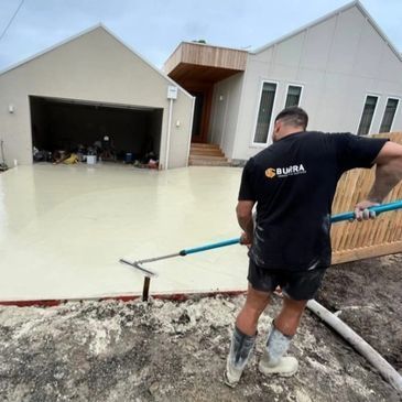 A man wearing work boots levels wet concrete on a driveway in front of a modern house.