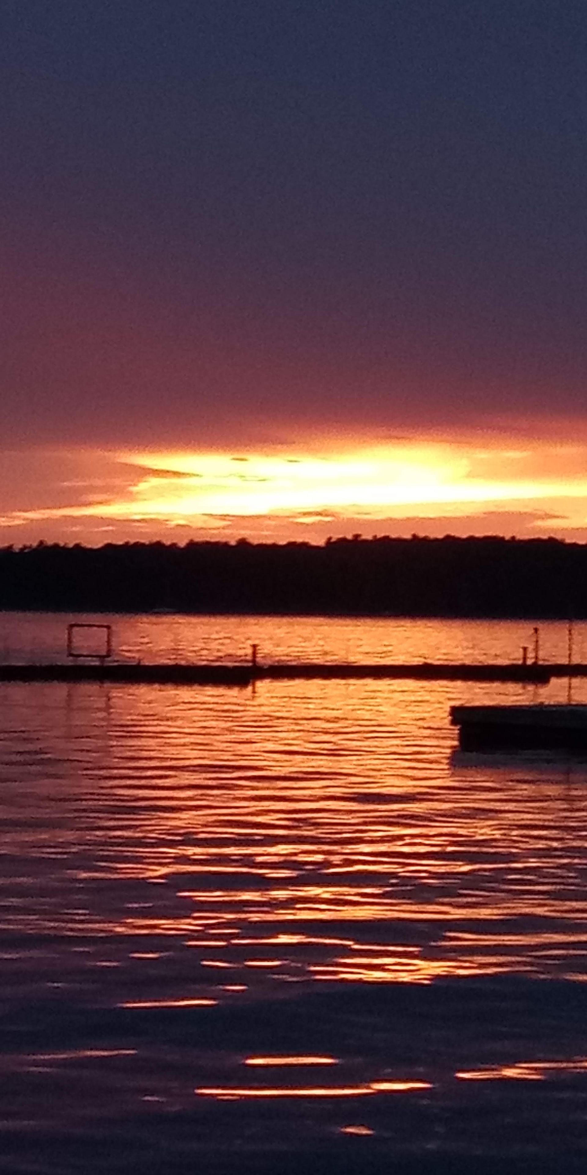 Sunset over a lake with a dock silhouetted against the colorful sky, reflecting in the water.