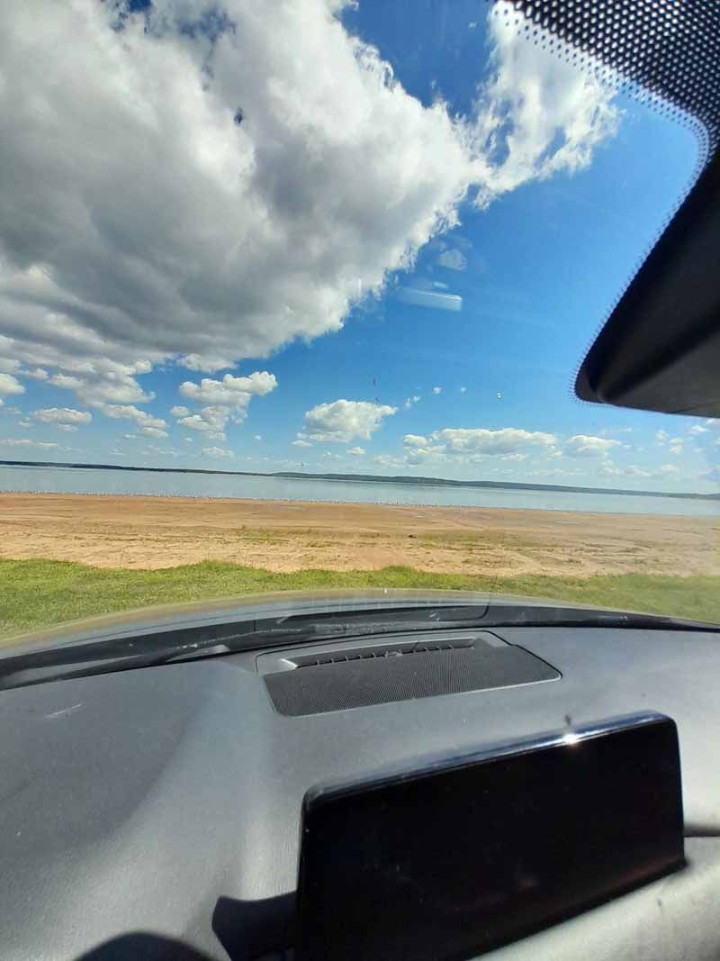 View from a car dashboard of a beach under a blue sky with puffy white clouds.