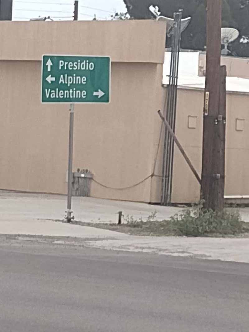 Green road sign pointing to Presidio, Alpine, and Valentine. Mounted on a pole near a beige building.