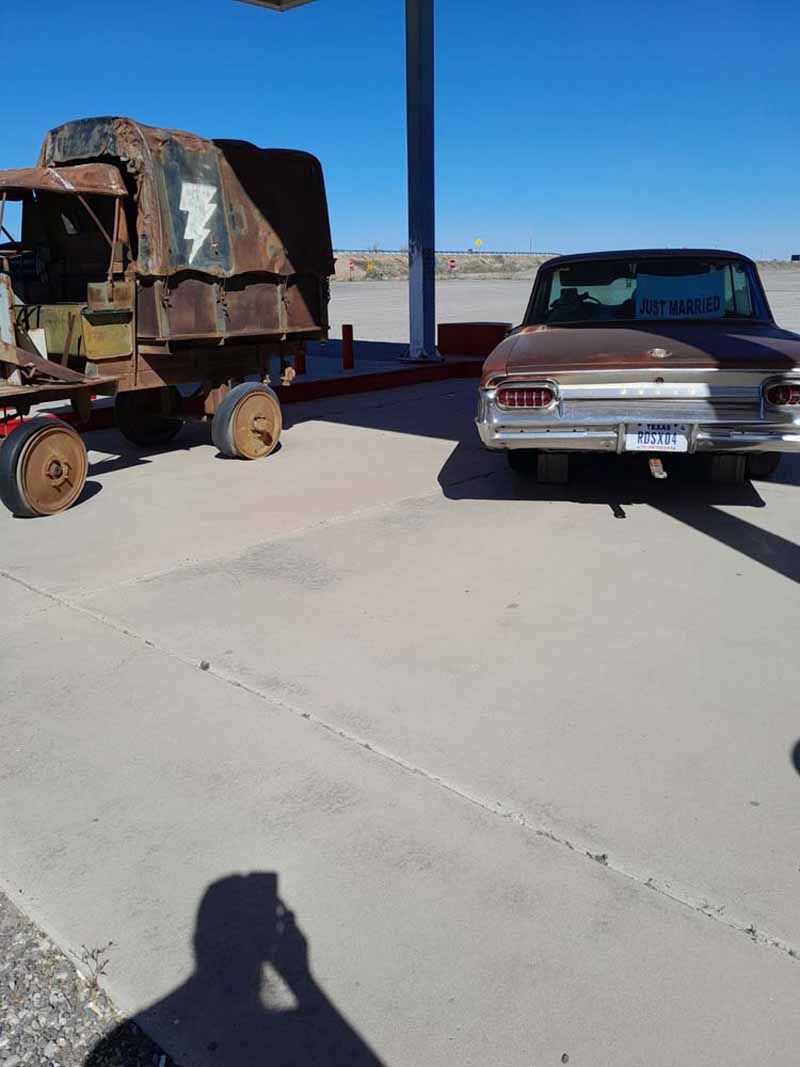 Vintage car and rusty trailer under a gas station canopy on a sunny day.