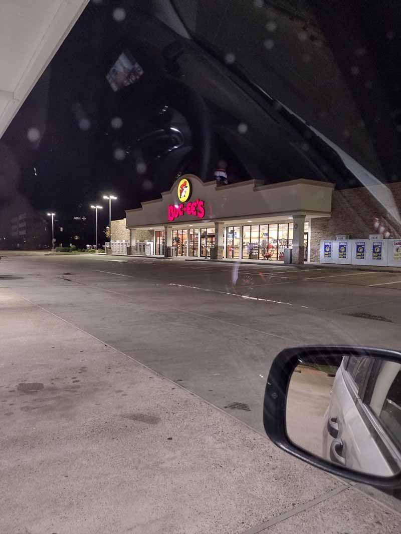 Nighttime view of a Buc-ee's gas station, illuminated with red signage. A side mirror of a car is in the frame.