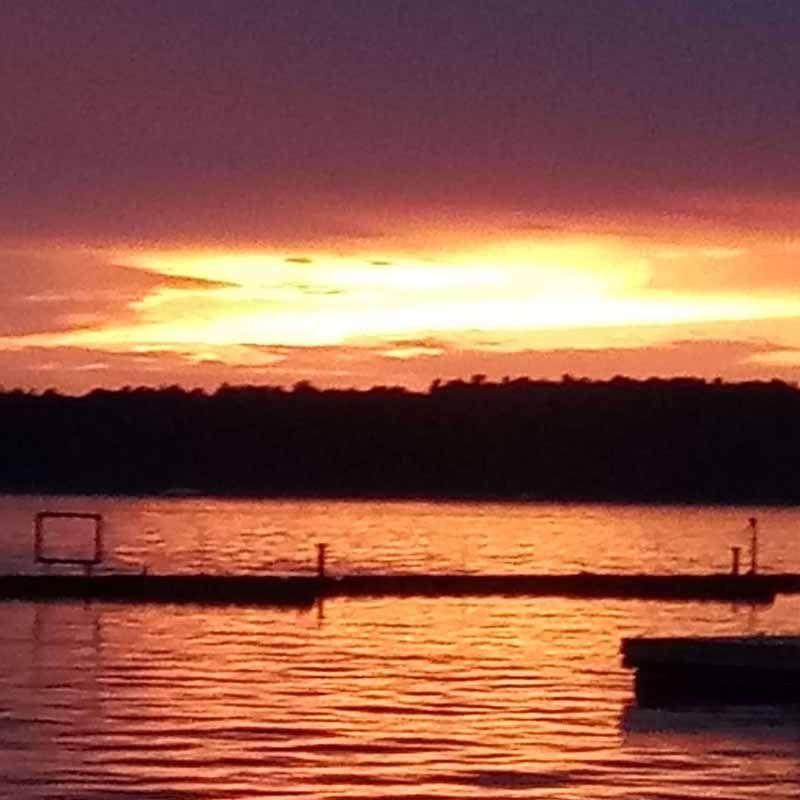Sunset over calm water with silhouetted docks and trees; orange and purple sky.