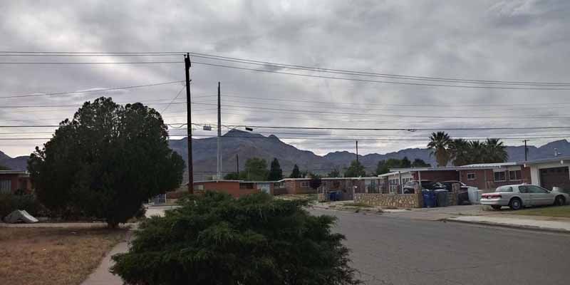 Street view with houses, trees, power lines, and mountains under a cloudy sky.