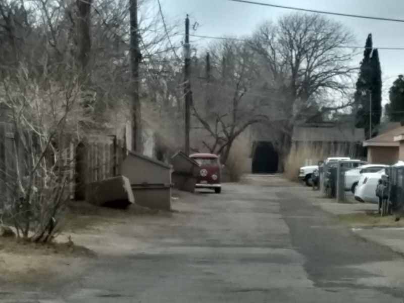 Alleyway with old red truck, cars parked on the right, tall trees, and a dark tunnel in the background.