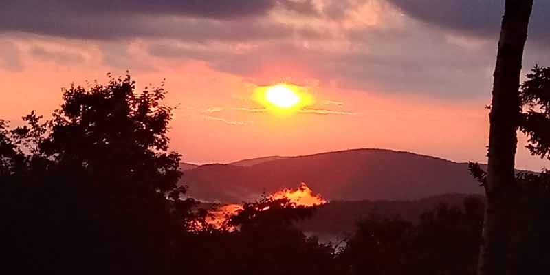 Sunset over mountains, with orange and pink hues in the sky. Silhouetted trees in the foreground.