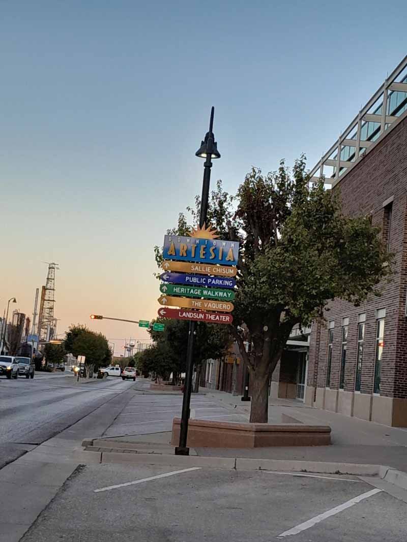 Street scene with signpost featuring multiple direction markers. Buildings line the street under a blue sky.