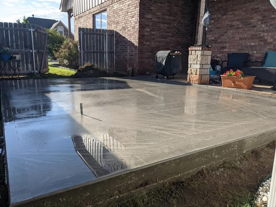 Concrete driveway leading to a house under construction, with a dark gray section and bordering dirt and trees.