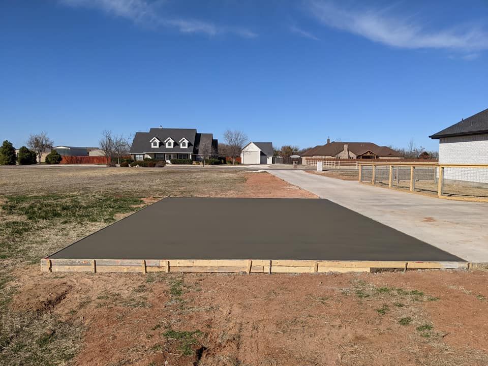 Newly poured concrete pad within wooden formwork, in a grassy field. Homes and a blue sky in the background.