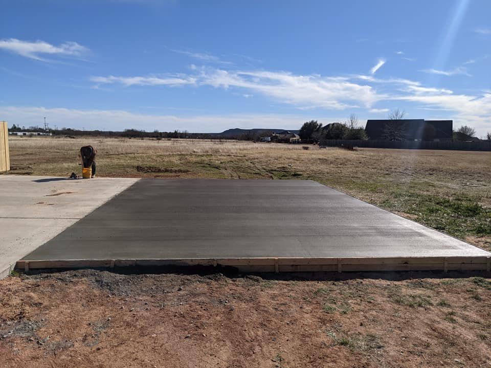 Freshly poured concrete slab in an open field, framed by wood, with person in the background, blue sky.