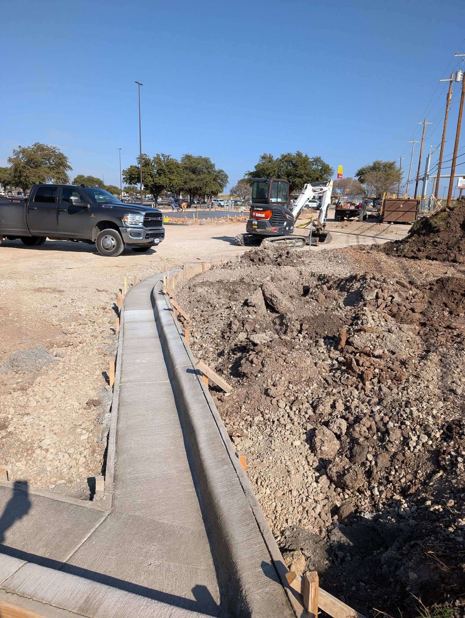 Construction site with truck, small excavator, and dirt pile; clear, sunny day.