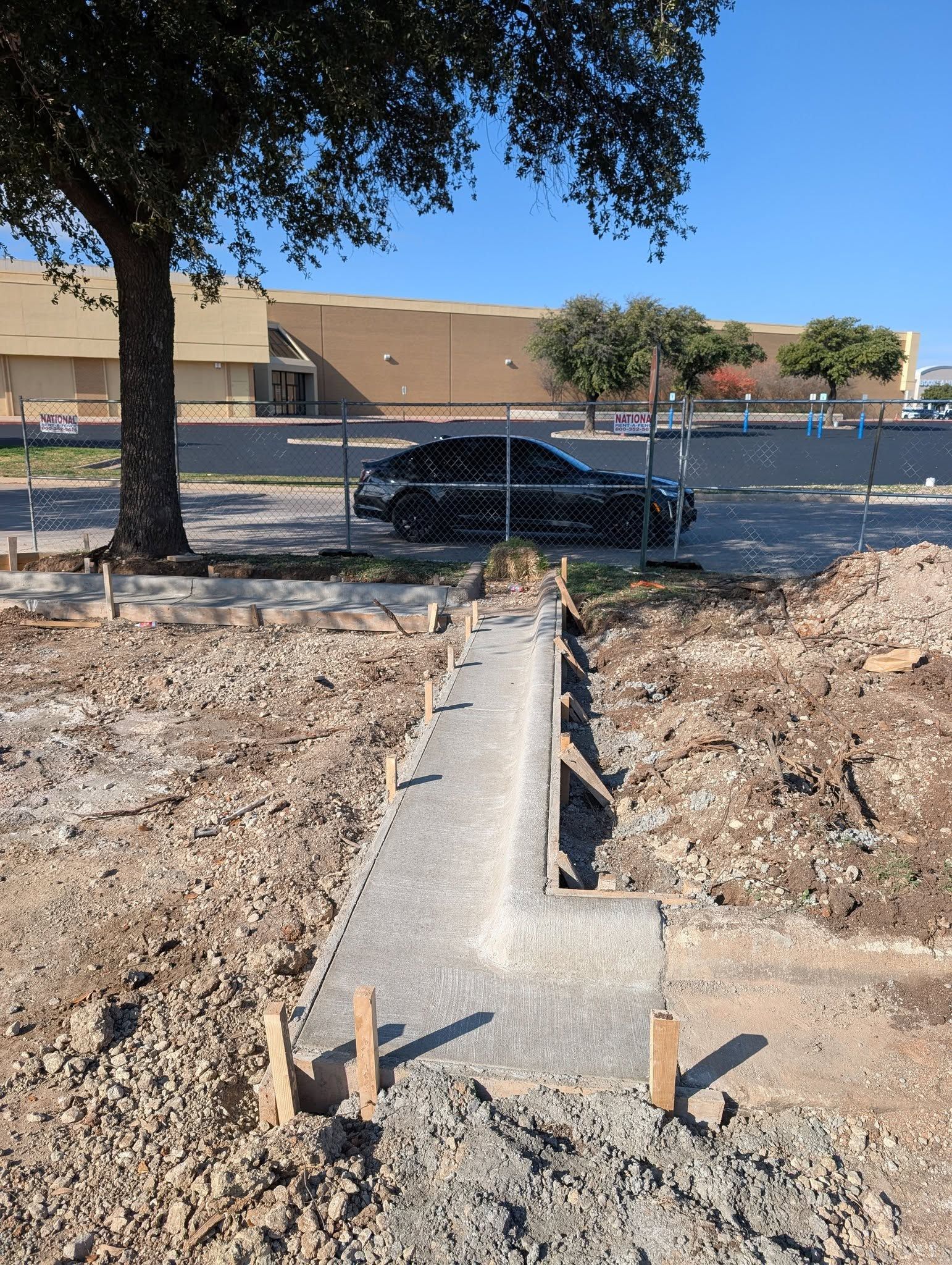 Construction site: concrete walkway under construction; dirt, gravel, and trees in the background.