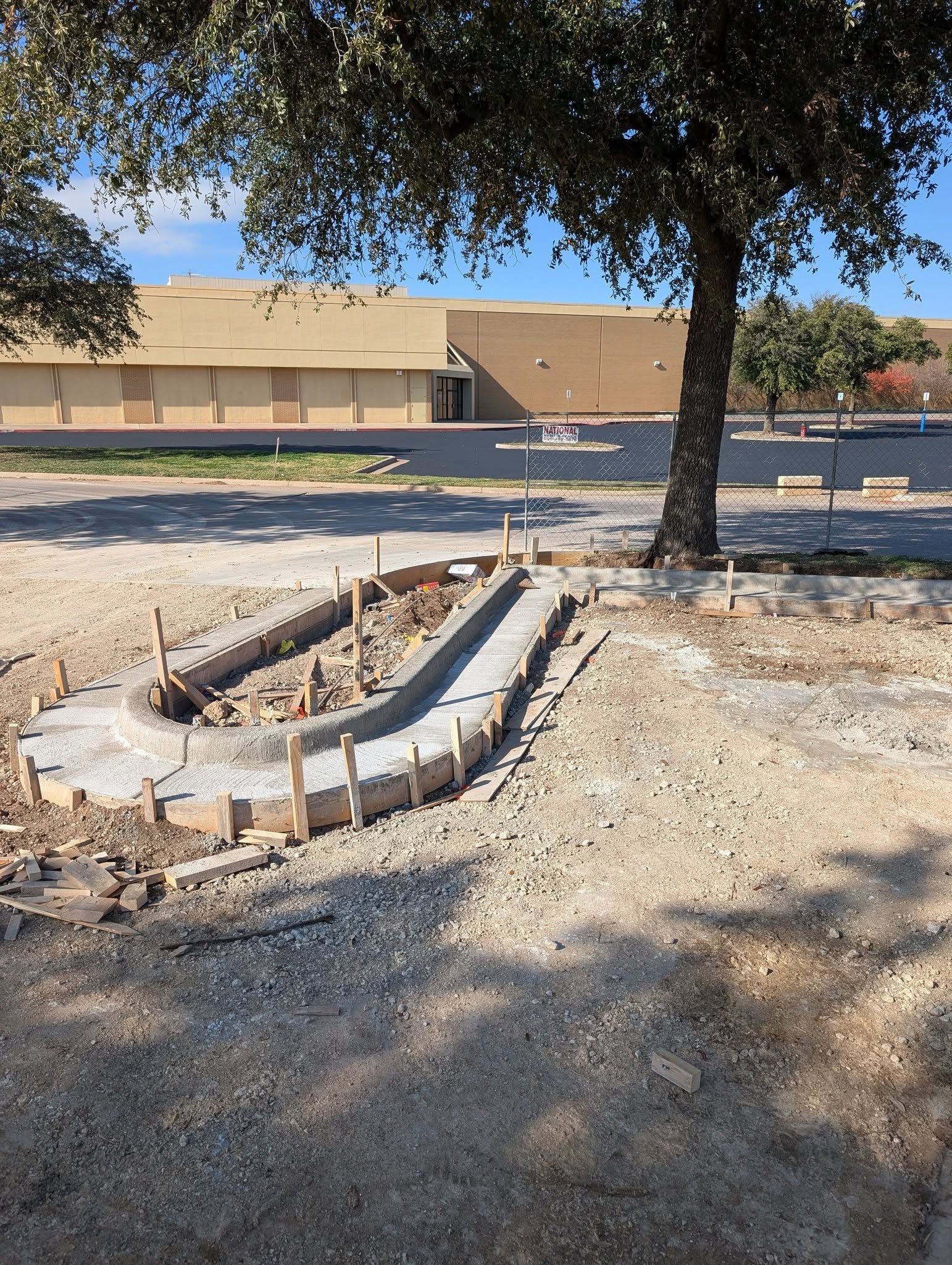 Construction site: Concrete forms for a curved pathway, gravel ground, tree, beige building in background.