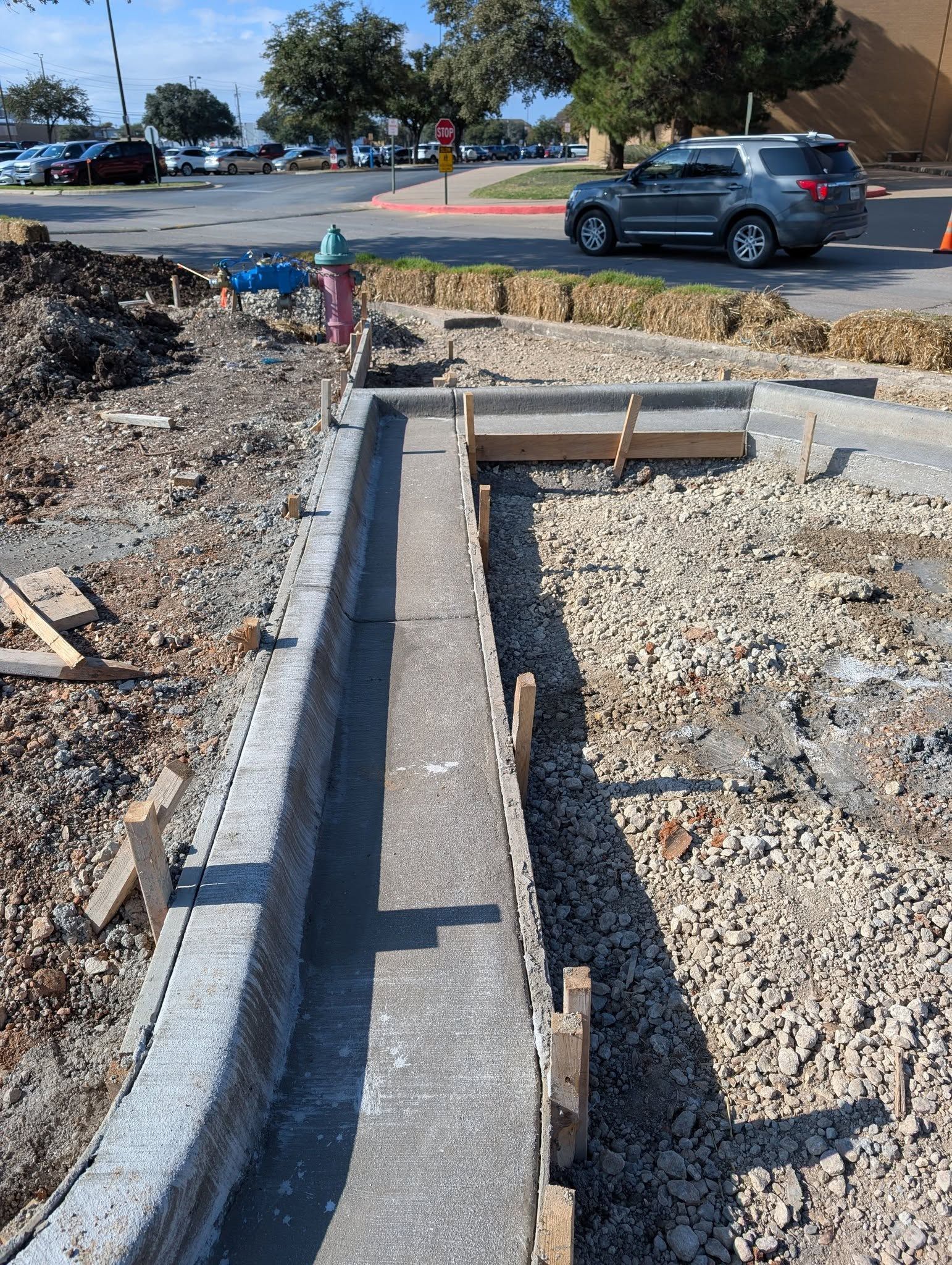 Sidewalk construction: gray concrete drainage channel, gravel, wooden stakes. Car and parking lot in background.