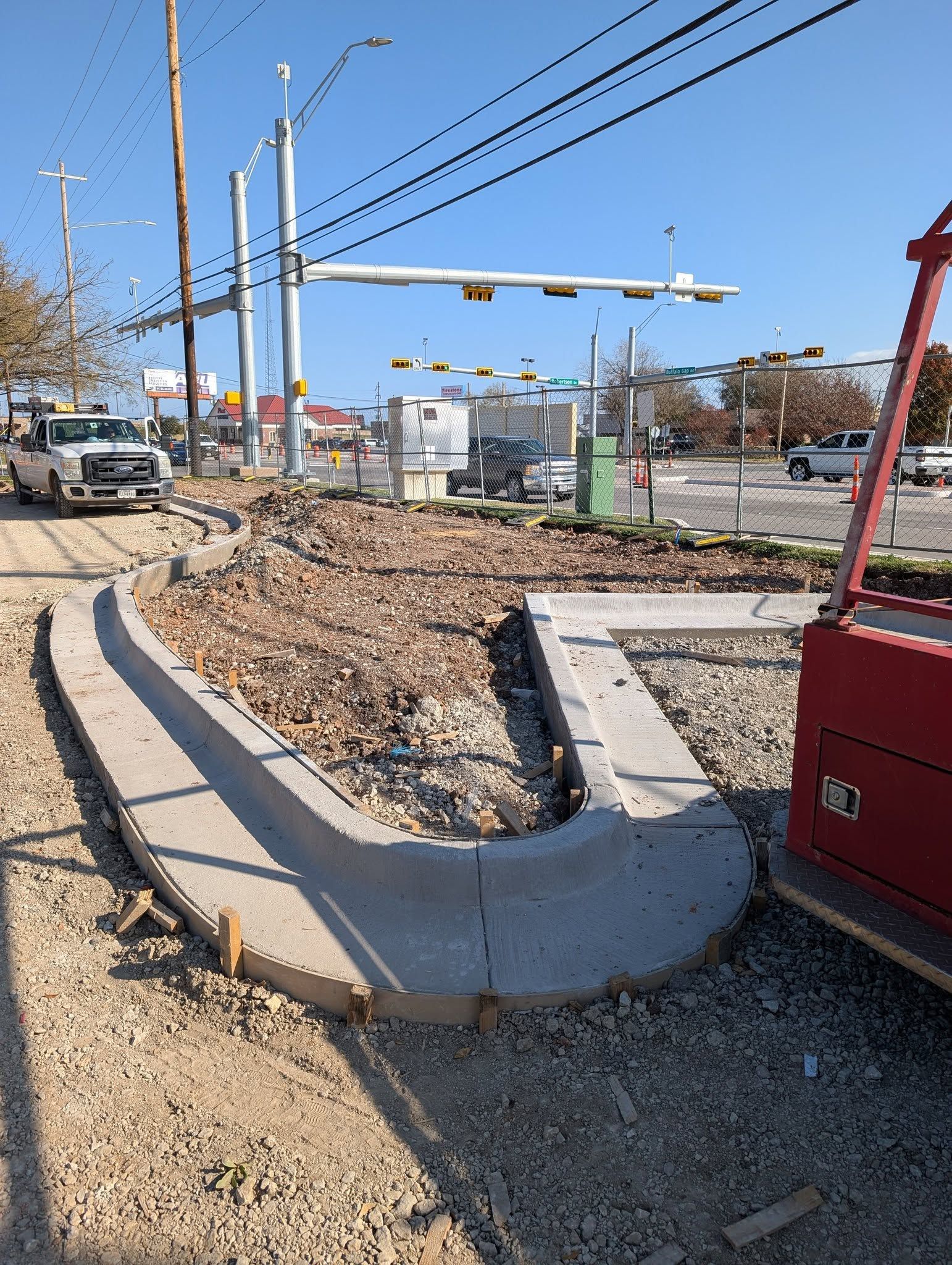 Curved concrete curb construction at a road intersection, with construction equipment and truck on a sunny day.