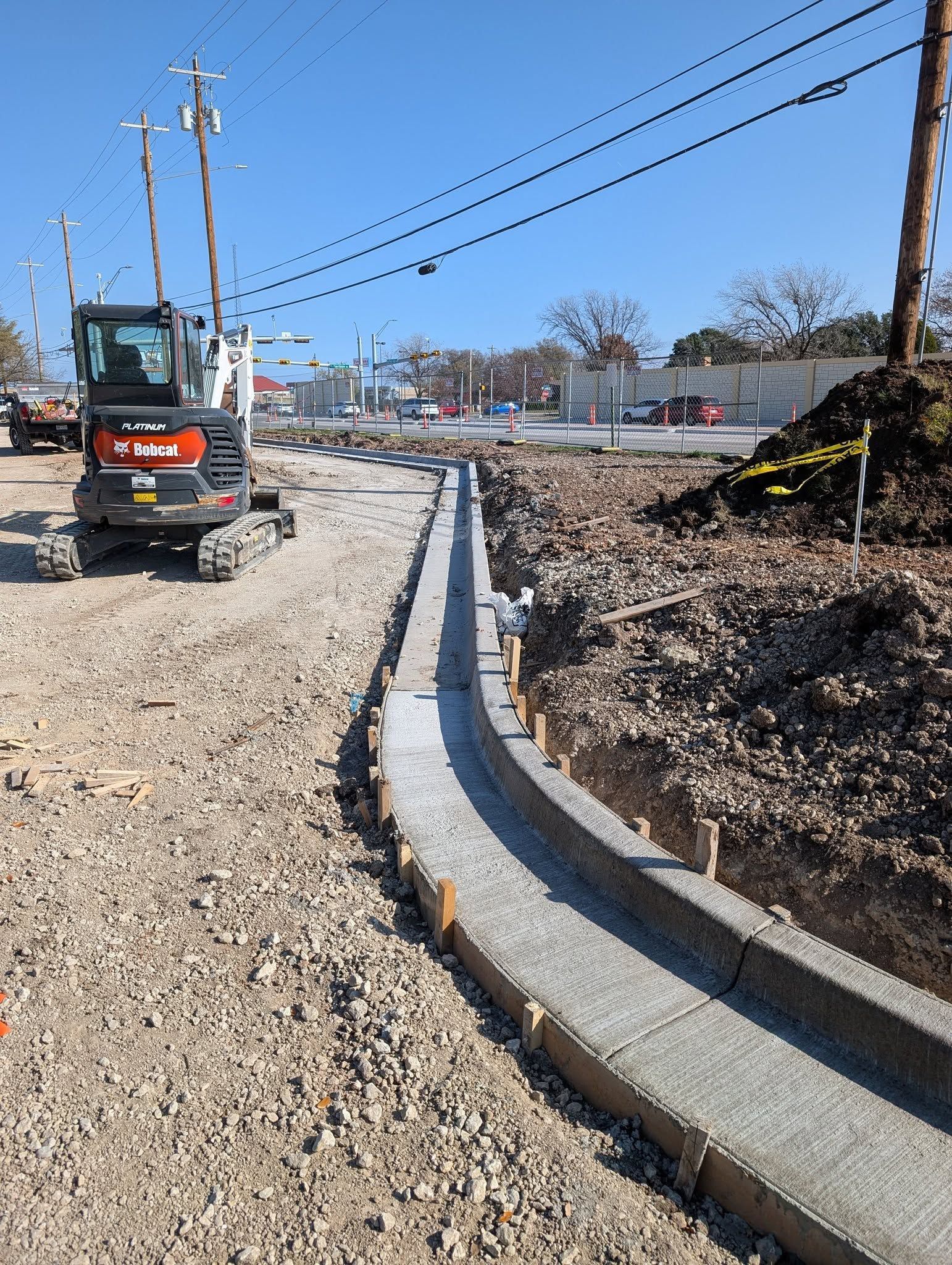 Construction site with a concrete channel being built, small excavator, and utility poles under a blue sky.