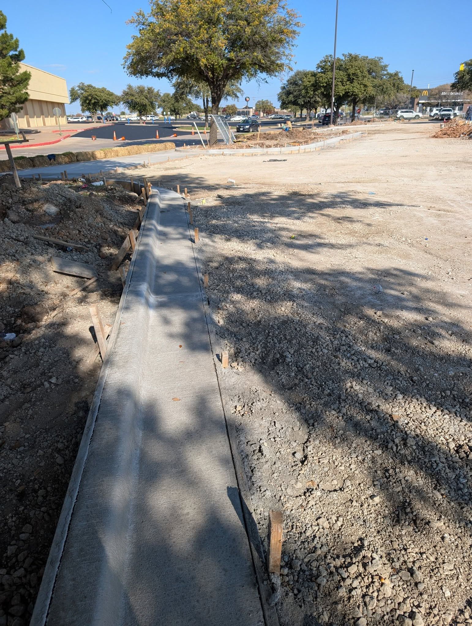 Concrete sidewalk under construction in a gravel lot, with trees and buildings in the background.