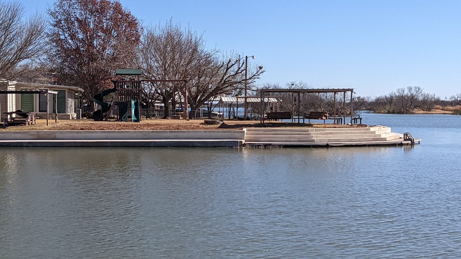 A boat ramp on a lake, with buildings and trees in the background, under a blue sky.