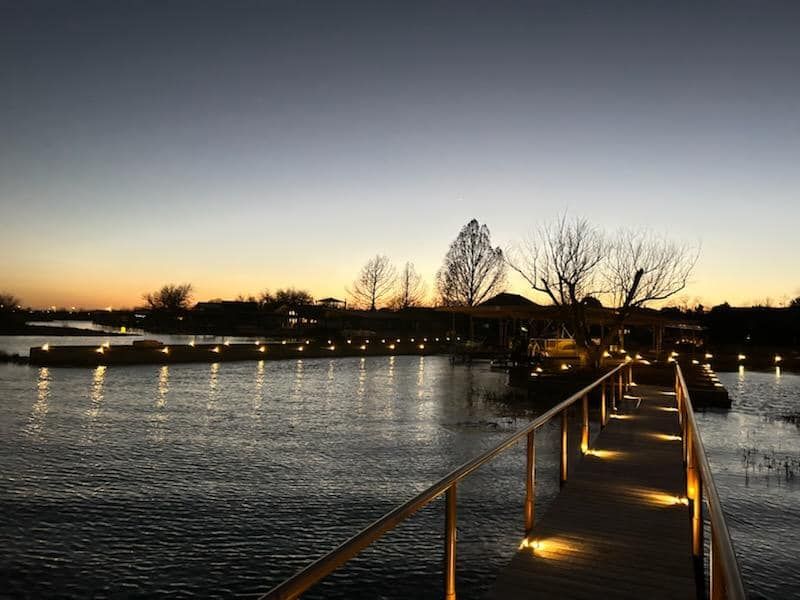 Wooden bridge over water, lit with warm lights, leads to a structure at dusk.