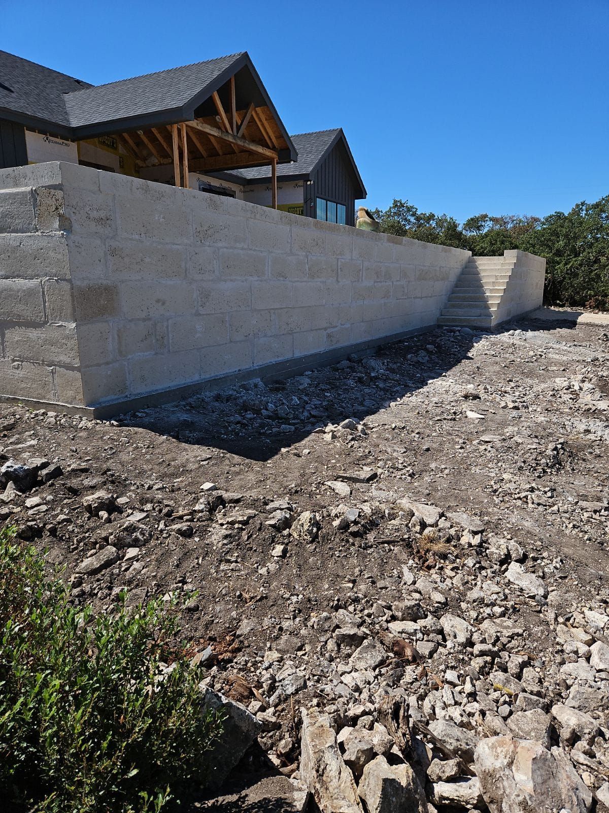 Beige stone retaining wall in front of a house under construction; dirt and rocks in foreground.