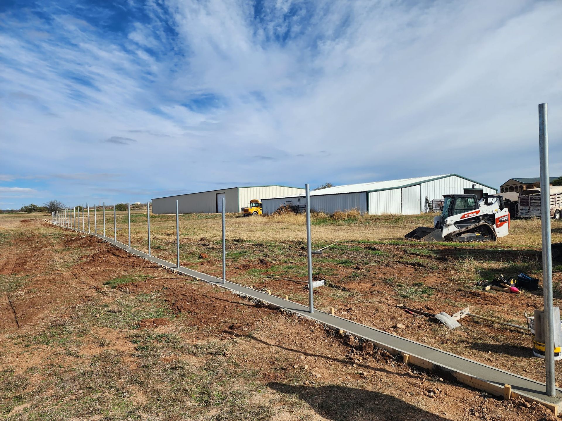 Construction site with a concrete foundation and metal fence posts in a rural area. White metal buildings in the background.