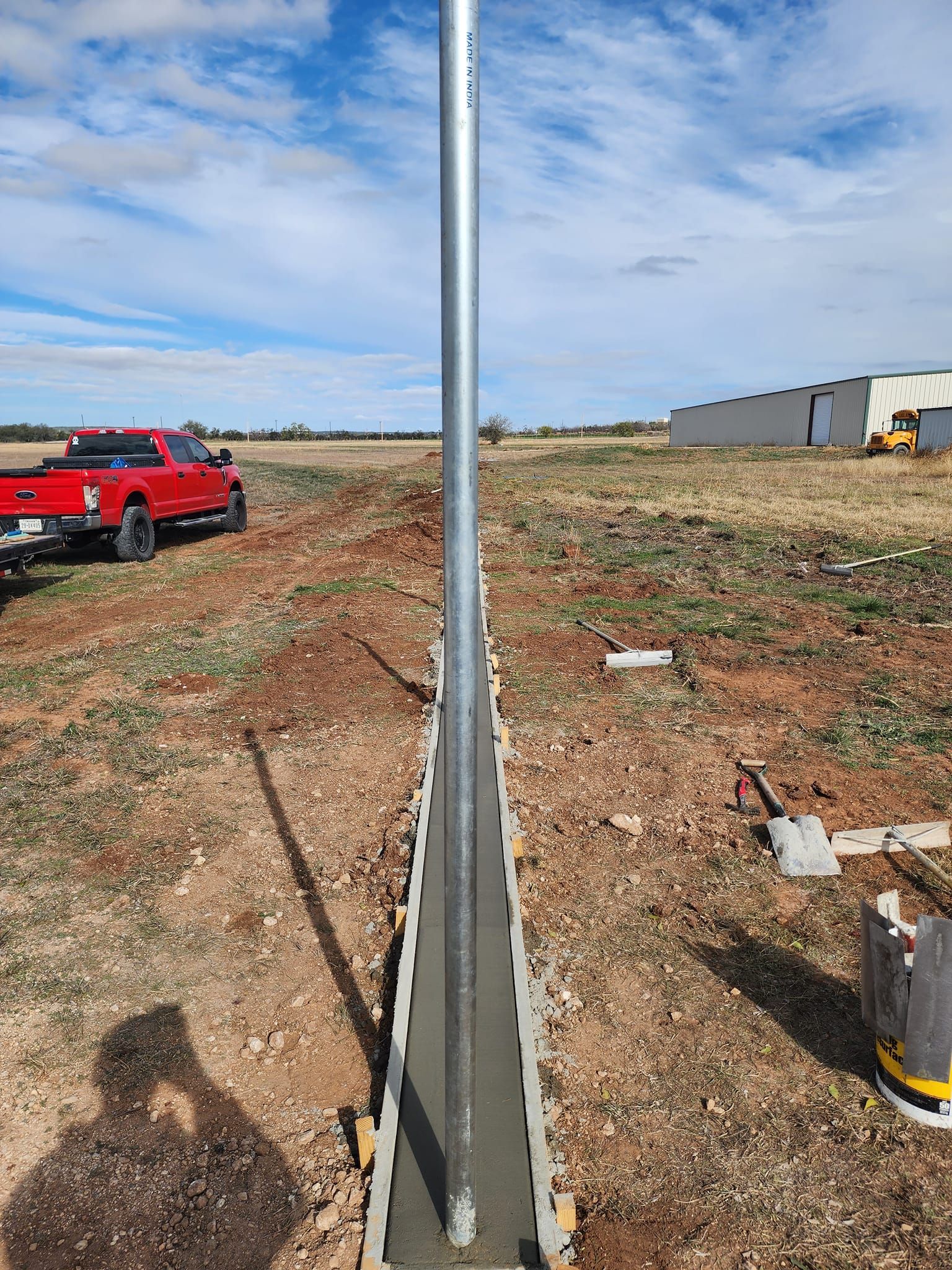 A metal pole set in fresh concrete trench, construction scene in field with red truck and building.