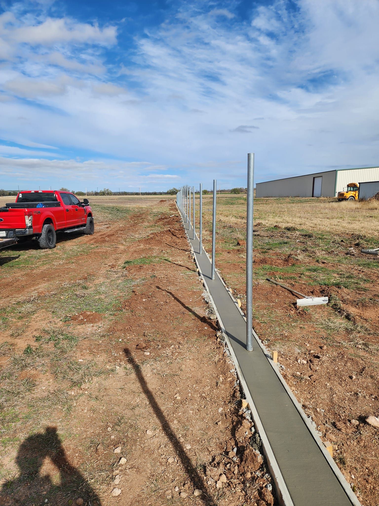 Fence construction with concrete base and metal posts; red truck on the left, industrial buildings in the background.