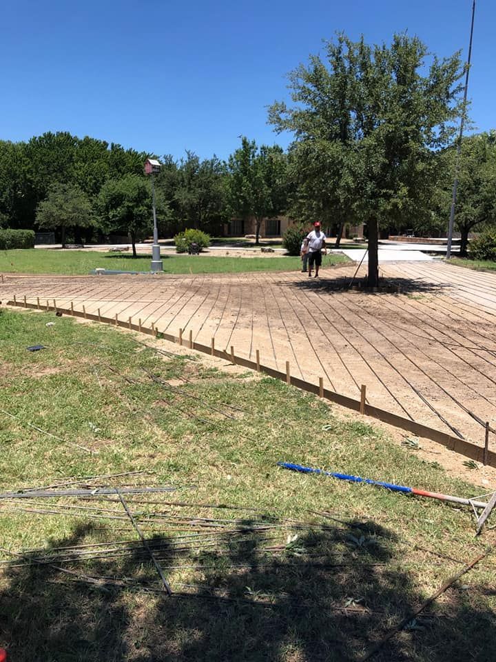 A person stands near a tree in a park. Concrete paving is in progress with rebar and forms.