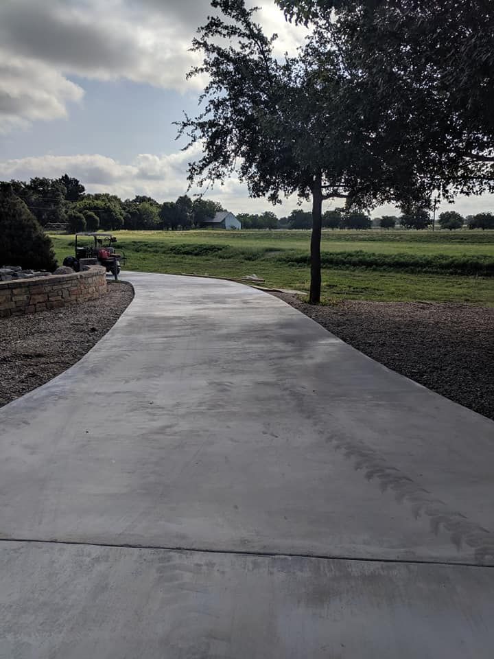 Concrete driveway curves towards a green field under a cloudy sky, with a tree on the right.