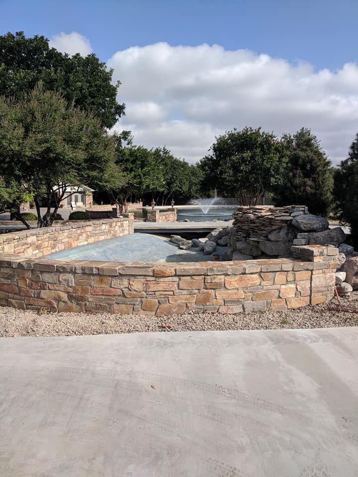 Stone-walled water feature with gravel and trees under a partly cloudy sky.
