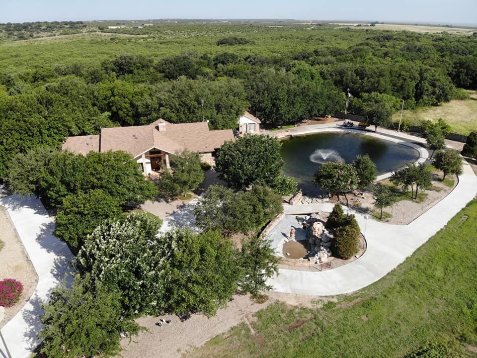 Large house nestled in trees by a pond, connected by a circular drive.
