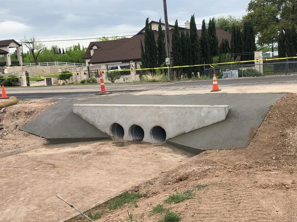 New concrete culvert with three openings, construction site, orange cones, and a house in the background.