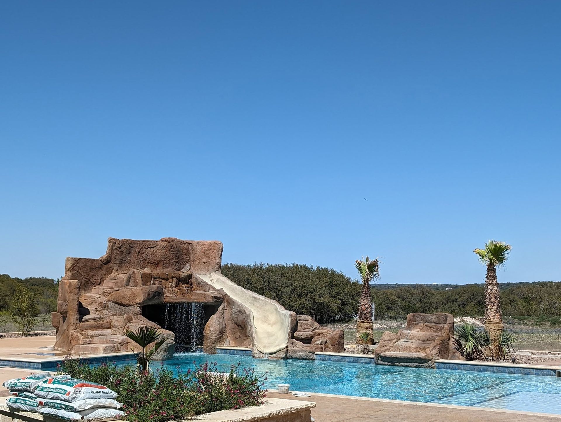 A swimming pool with a rock waterfall, slide, and palm trees under a blue sky.