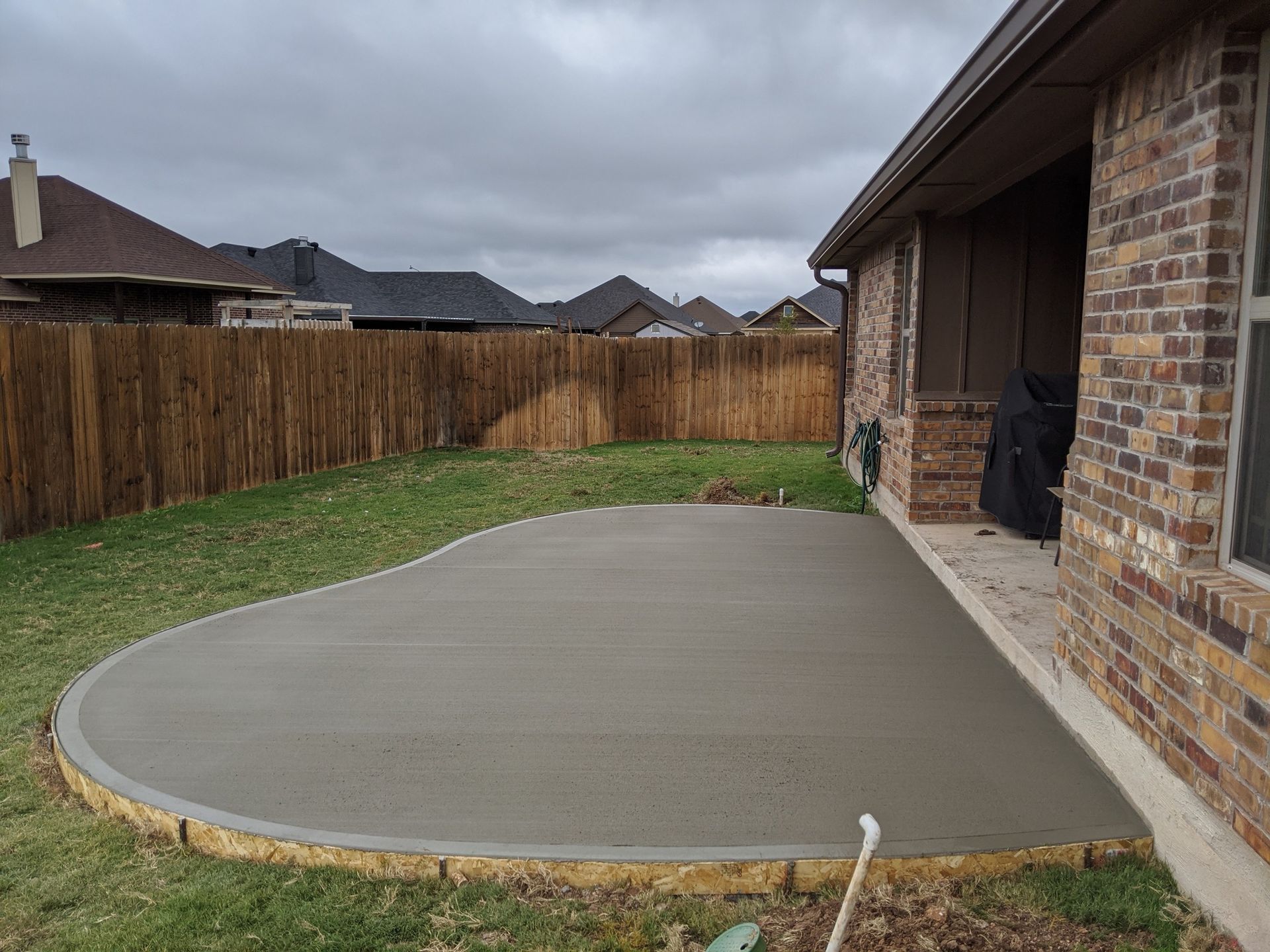 Newly poured concrete patio, curved edge, next to a brick house, green yard, brown fence.