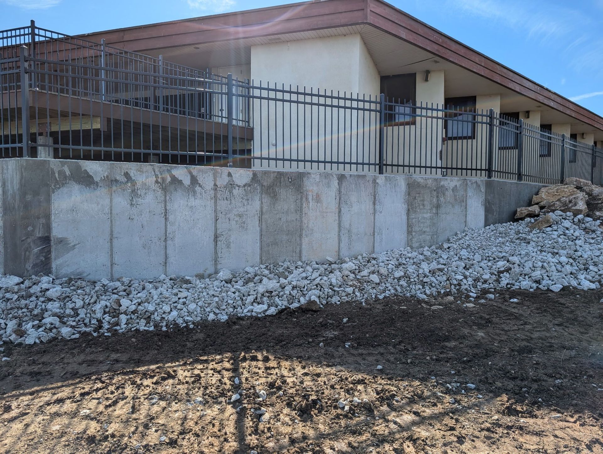 Building with a concrete retaining wall, metal fence, and gravel slope. Brown and tan tones, outdoors.