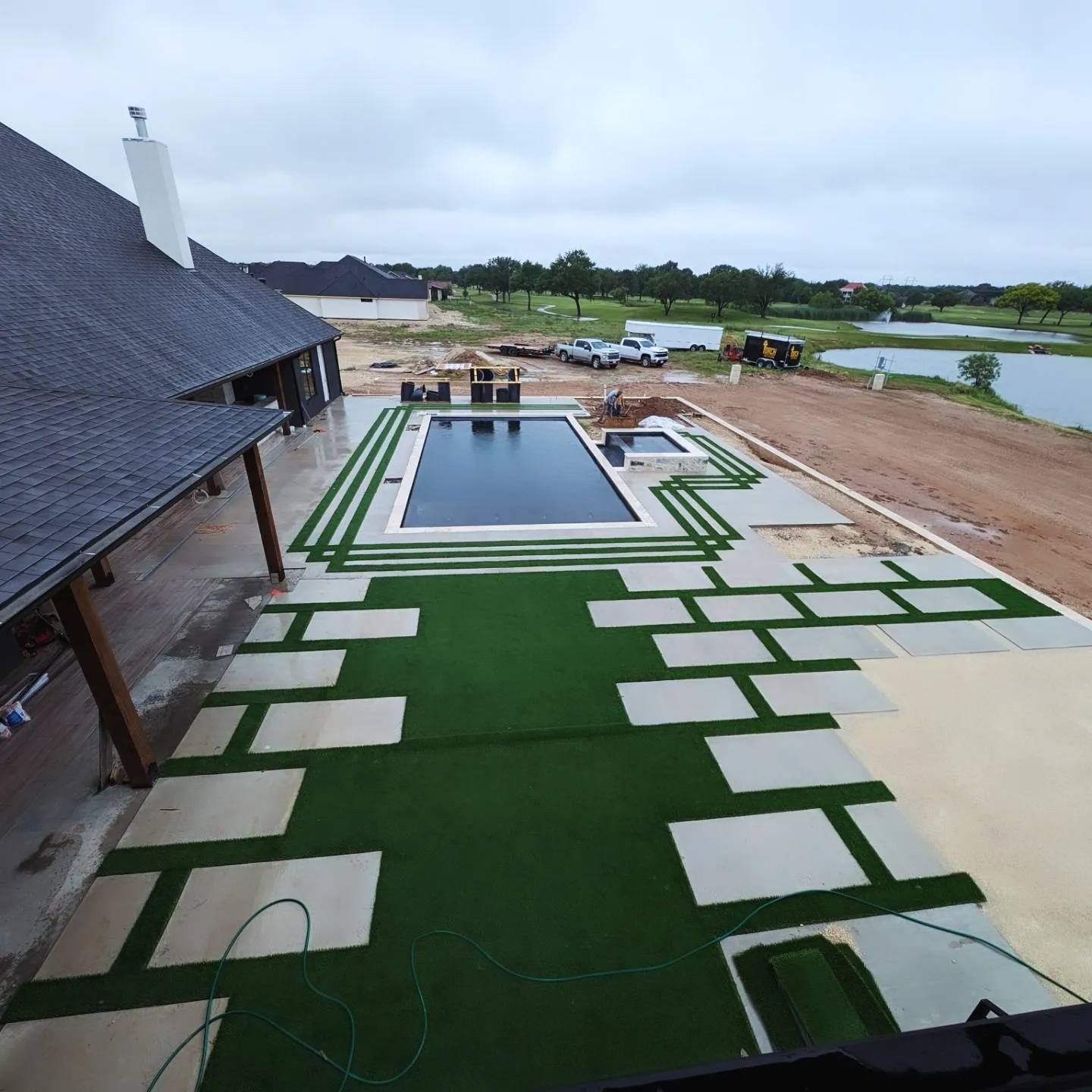 Backyard with pool, concrete pavers, and artificial turf near a lake under overcast skies.